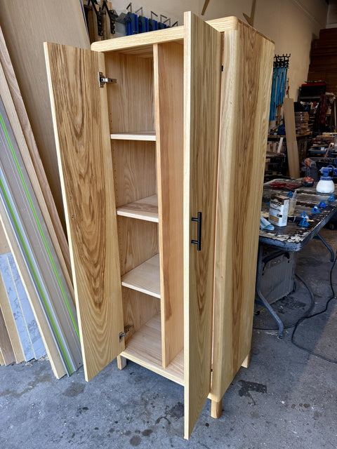 A tall, narrow wooden cabinet with two doors, shown open to reveal four interior shelves. The cabinet is made of light natural wood with black metal handles and hinges, photographed in a workshop environment.