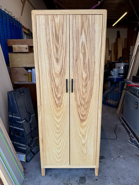 A tall wooden two-door cabinet with a natural ash finish and black metal handles