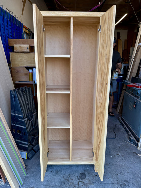 A tall, light natural wood storage cabinet with two open doors and four interior shelves, photographed in a workshop environment.