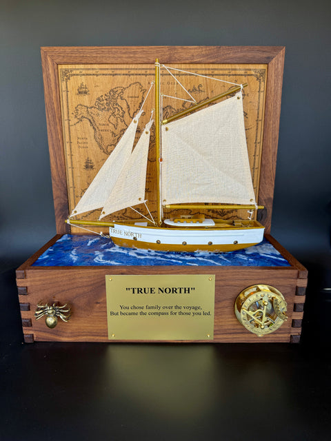 A wooden model nautical display featuring a detailed sailing ship with cream sails mounted on a blue wave-patterned base inside a wooden case. The front has a brass plaque reading "TRUE NORTH" and a brass compass ornament, set against a dark background.
